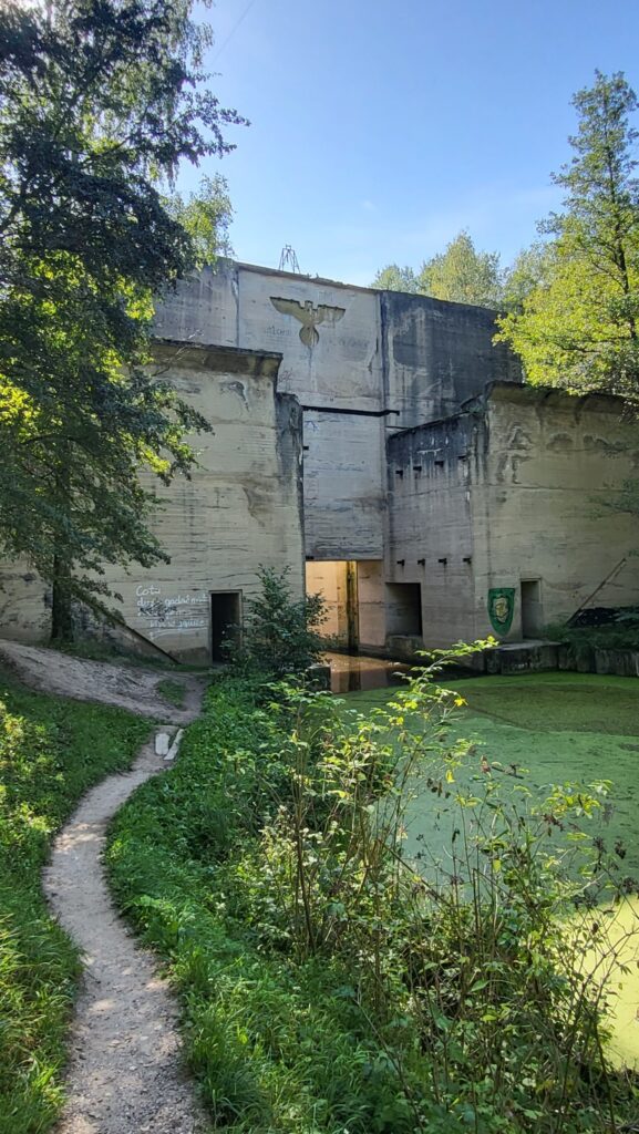 Wejście do historycznego bunkra na trasie Mazurskiej Pętli Rowerowej, otoczone zielenią i malowniczą ścieżką prowadzącą przez las.