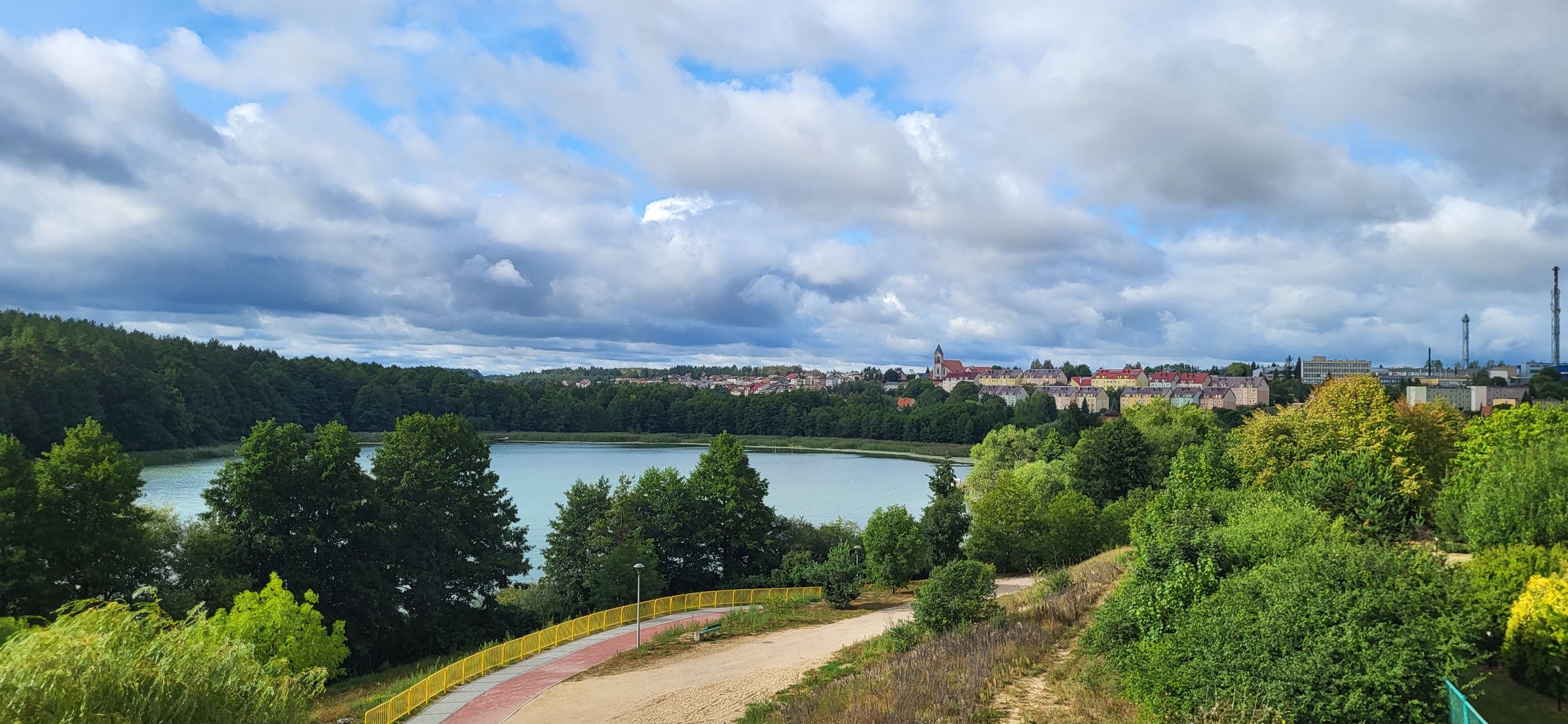 Widok na malownicze Mazury z perspektywy Mazurskiej Pętli Rowerowej. Na pierwszym planie widać krętą ścieżkę rowerową z żółtymi barierkami, otoczoną bujną zielenią. W tle rozciąga się spokojne jezioro, za którym wznosi się urokliwa panorama miasta z kolorowymi dachami i wieżą kościoła. Niebo zdobią dynamiczne chmury, dodając krajobrazowi niezwykłego uroku. Idealne miejsce na relaks i rowerową eksplorację natury oraz lokalnej architektury.